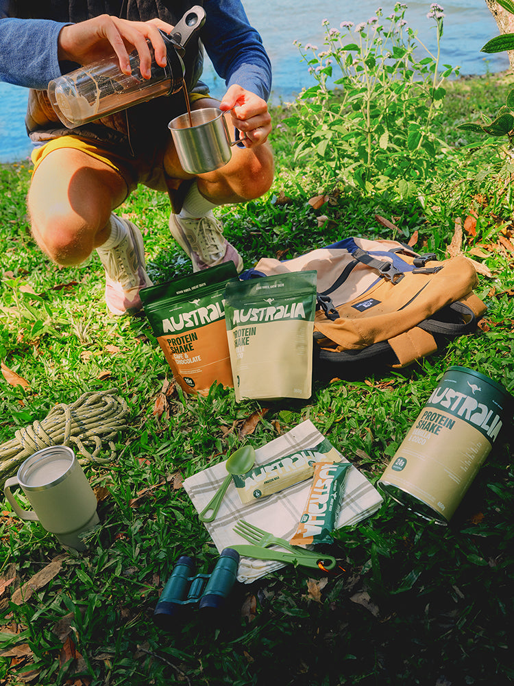 Pessoa prepara um shake em uma mesa de acampamento ao ar livre, com embalagens AUSTRALIA ao lado.