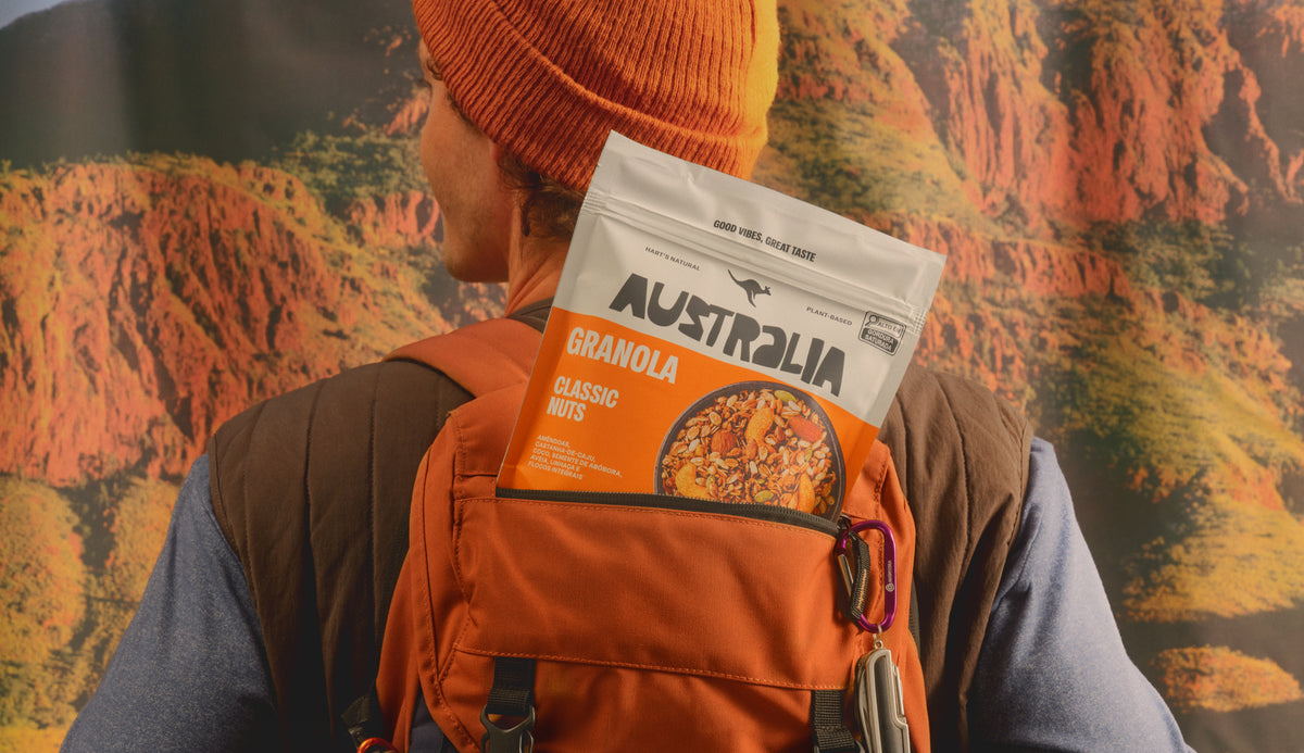 Pessoa com uma mochila levando uma embalagem de Granola Classic Nuts AUSTRALIA diante de uma paisagem de montanha.