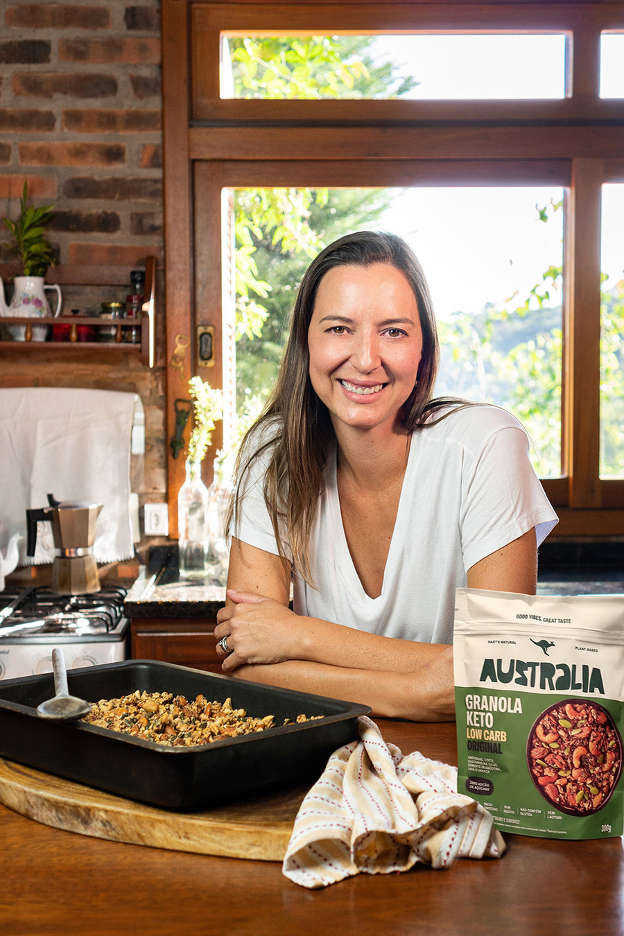 Raquel despeja leite de uma jarra numa tigela de granola na bancada da cozinha, com um pacote de Granola Keto Original ao lado.