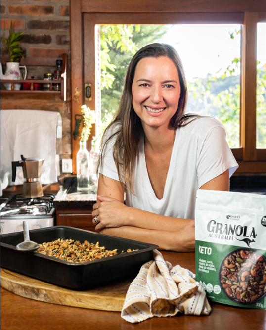 Raquel despeja leite de uma jarra numa tigela de granola na bancada da cozinha, com um pacote de Granola Keto Original ao lado.