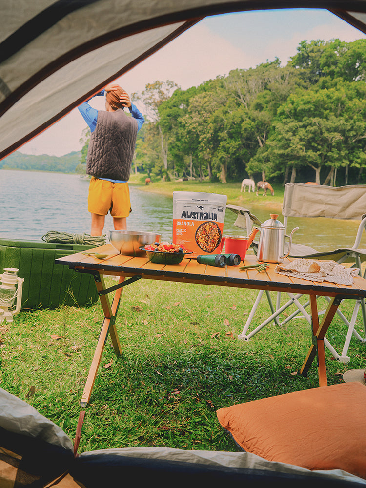 Mesa de acampamento com embalagens de granola AUSTRALIA e uma pessoa ao fundo.
