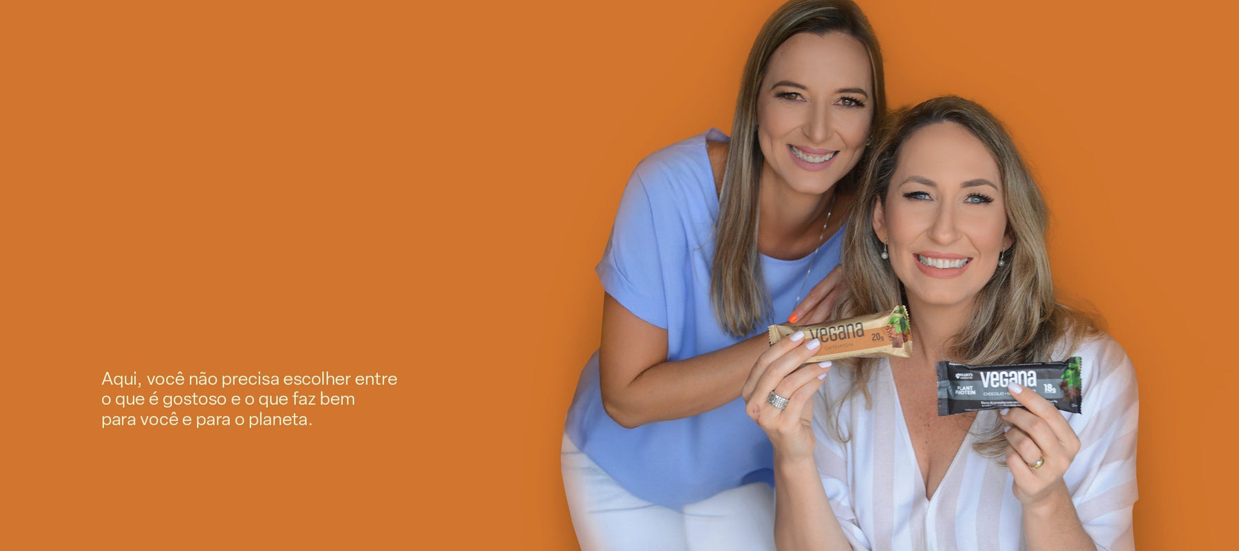 Duas mulheres sorrindo e segurando barras de proteína diante de um fundo laranja.