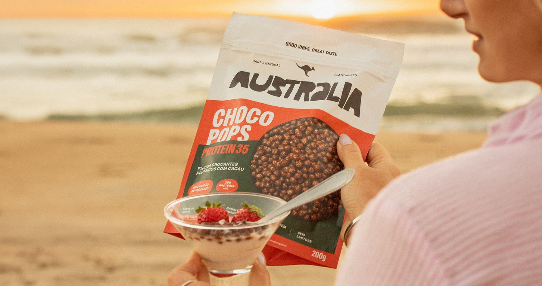 Pessoa segura uma embalagem de ChocoPops Protein e uma tigela com frutas à beira-mar.