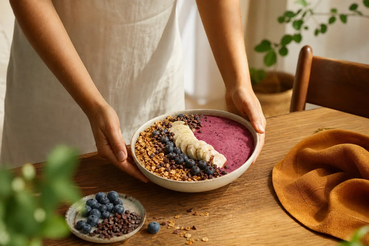 Pessoa segura uma tigela de vitamina grossa com banana, mirtilo, cacau e granola.
