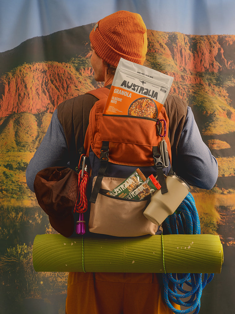 Pessoa de costas com mochila, uma granola e duas barras da AUSTRALIA, diante de uma paisagem de montanha.
