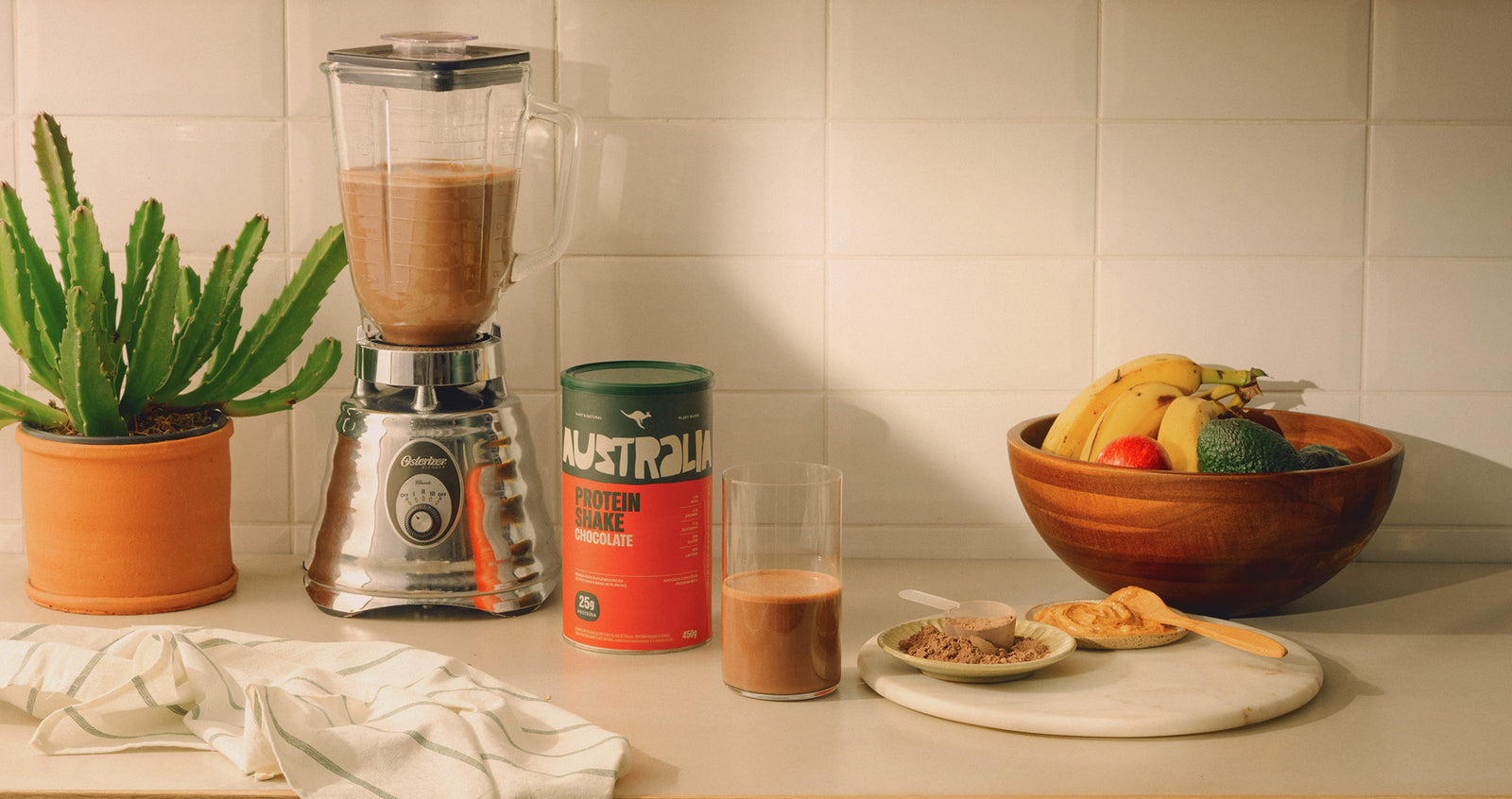 Protein Shake Chocolate 450 g ao lado do liquidificador e de um copo com shake em uma bancada de cozinha.