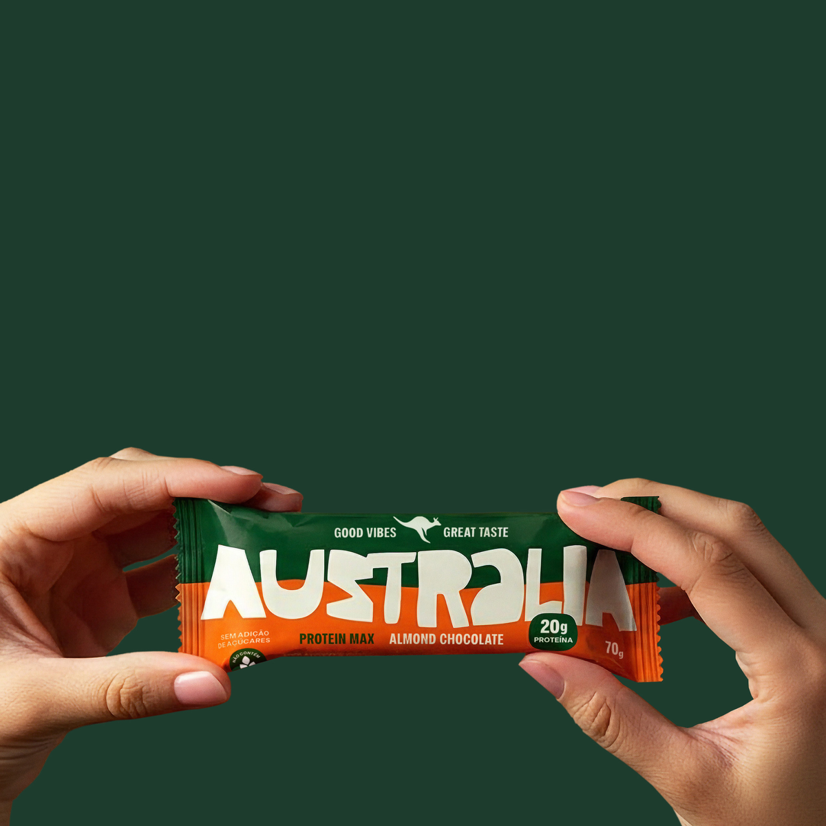 Duas mãos seguram uma barra Protein Max Amêndoa & Chocolate sobre fundo verde-escuro.