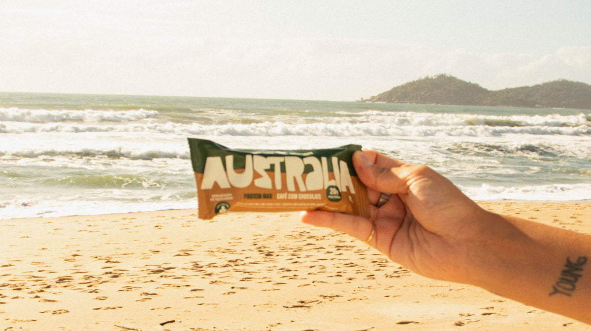 Mão segura uma barra Protein Max Café & Chocolate na praia, com o mar ao fundo.