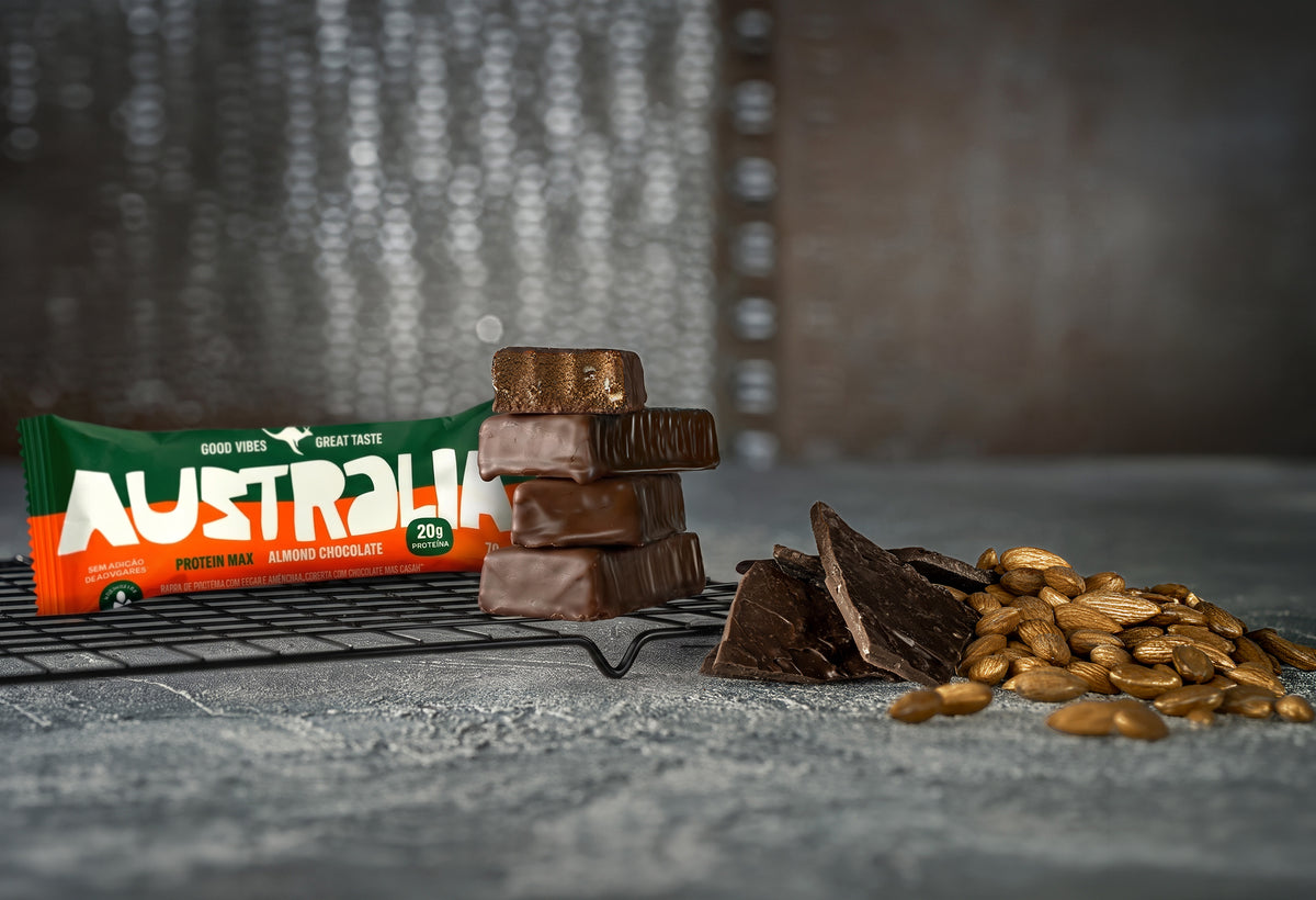 Uma Protein Max Amêndoa & Chocolate fechada sobre uma grade, ao lado de pedaços da barra empilhados, com lascas de chocolate e amêndoas na bancada.