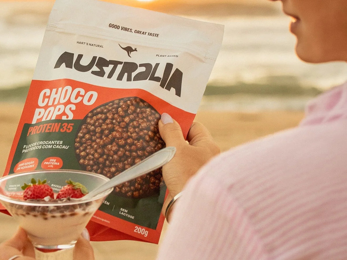 Mão segurando o pacote de ChocoPops Protein 35 e uma taça com cereal e iogurte